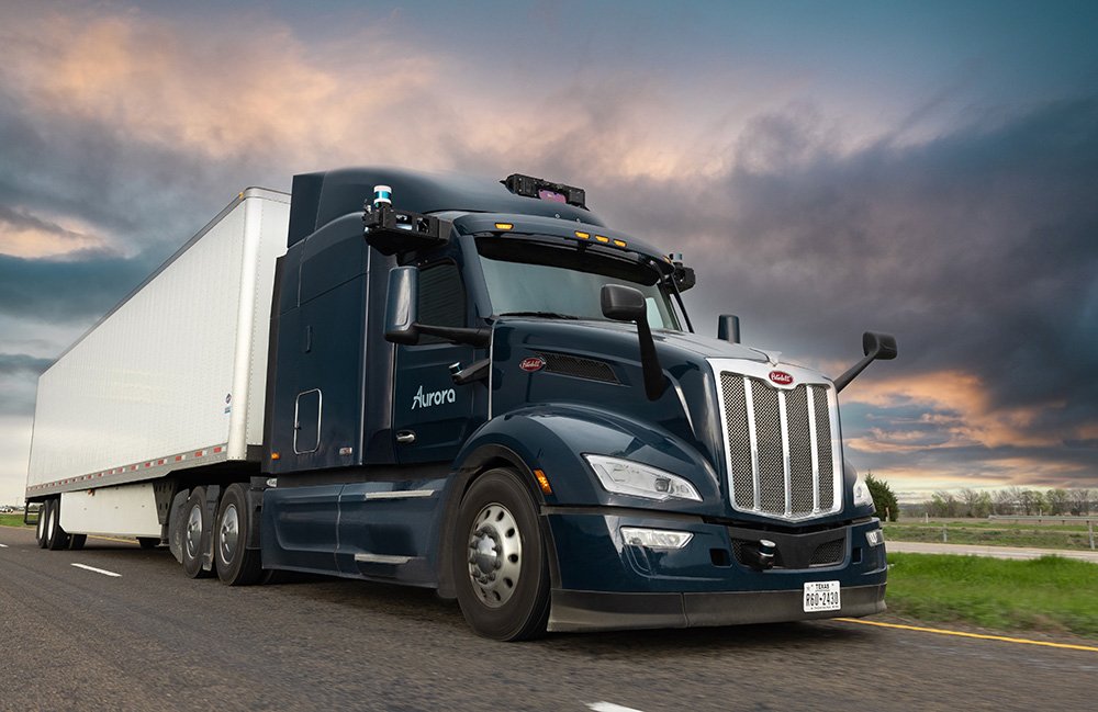 An Aurora autonomous truck driving on a road with a sunset in the background.